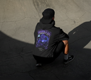 Person wearing a black hoodie with a graphic design on the back, sitting on a cracked pavement.