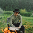Man cooking over an open fire in a natural setting with trees and water in the background.